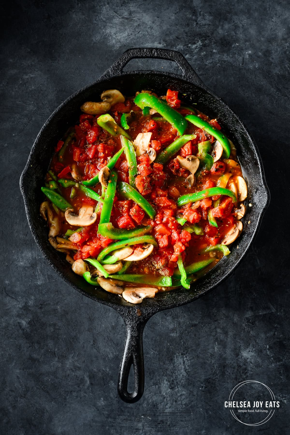 Overhead view of bell peppers, mushrooms, garlic, oregano, and diced tomatoes in a cast iron skillet