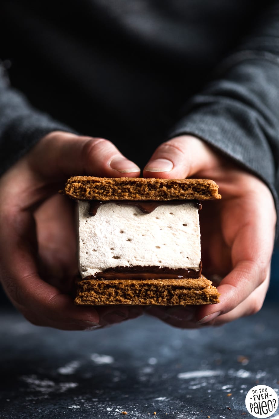 Hands holding a paleo s'mores bar with graham crackers, melted chocolate, and marshmallow