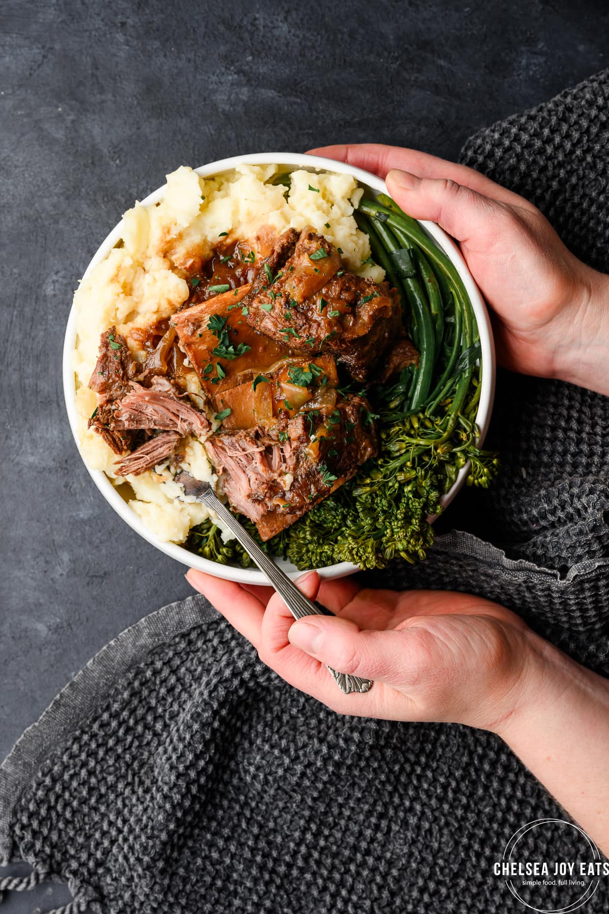 Hands holding a bowl of apple cider braised short ribs over mashed potatoes and roasted broccolini on a dark background