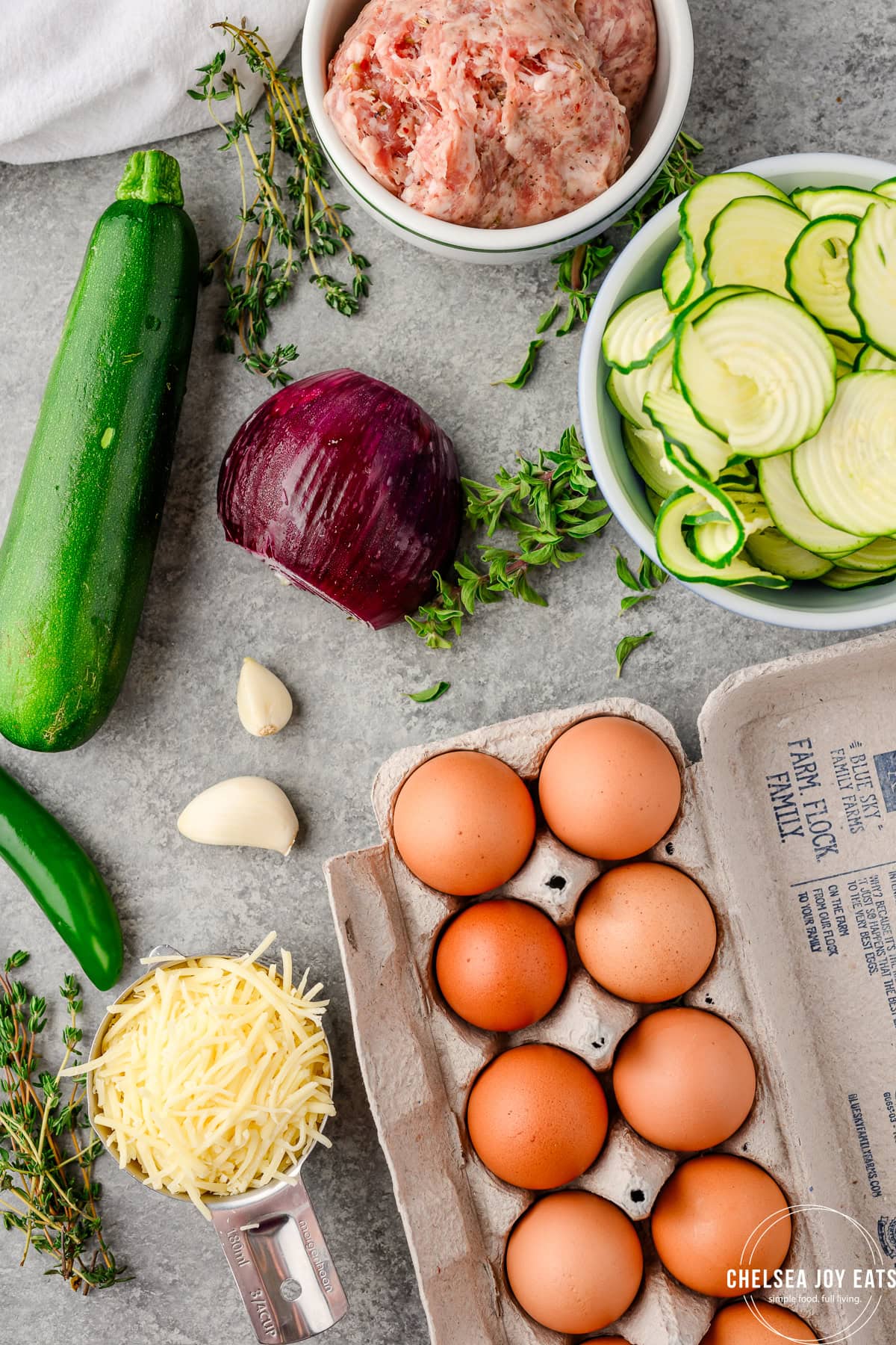 Ingredients for a breakfast casserole, including eggs, zucchini, onion, sausage, herbs, and cheese