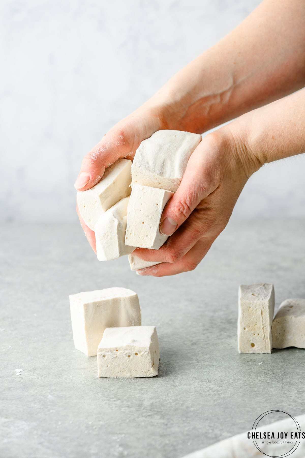 Hands holding multiple homemade marshmallows with more marshmallows on the table below