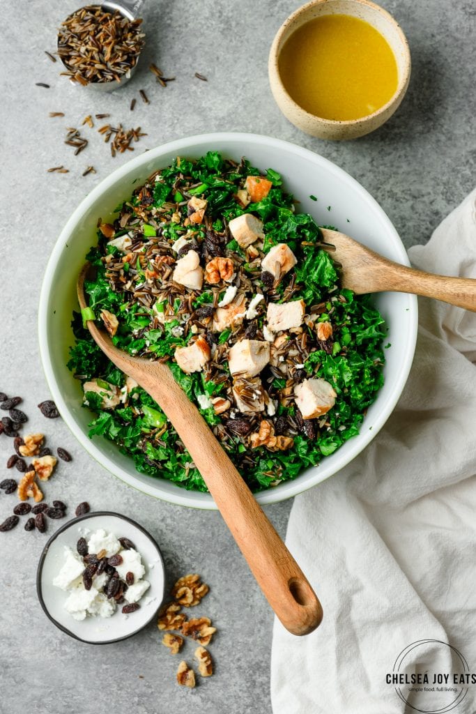 Overhead view of a mixing bowl with wild rice salad ingredients, including kale and chicken with orange dressing on the side