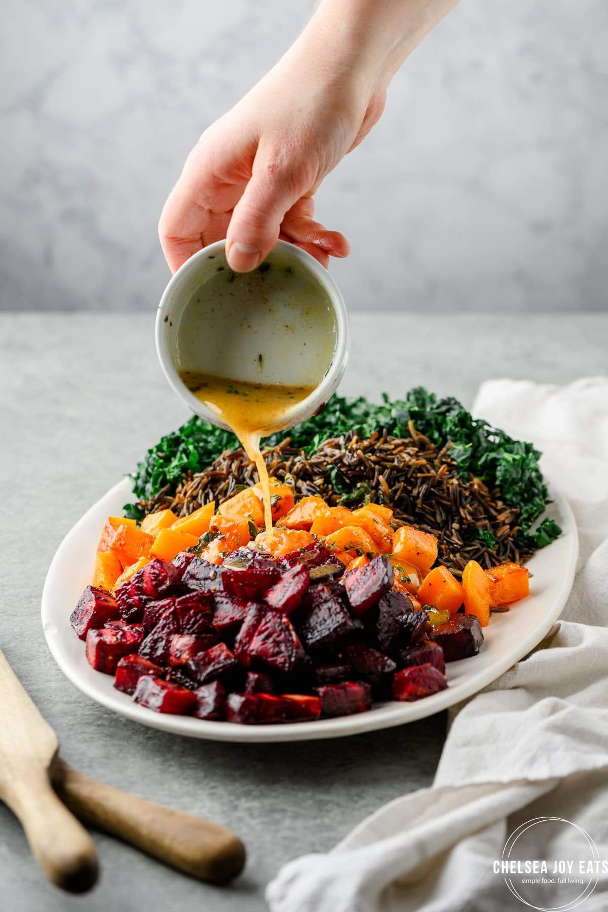 Pouring dressing over a platter with roasted beets, butternut squash, kale, and wild rice