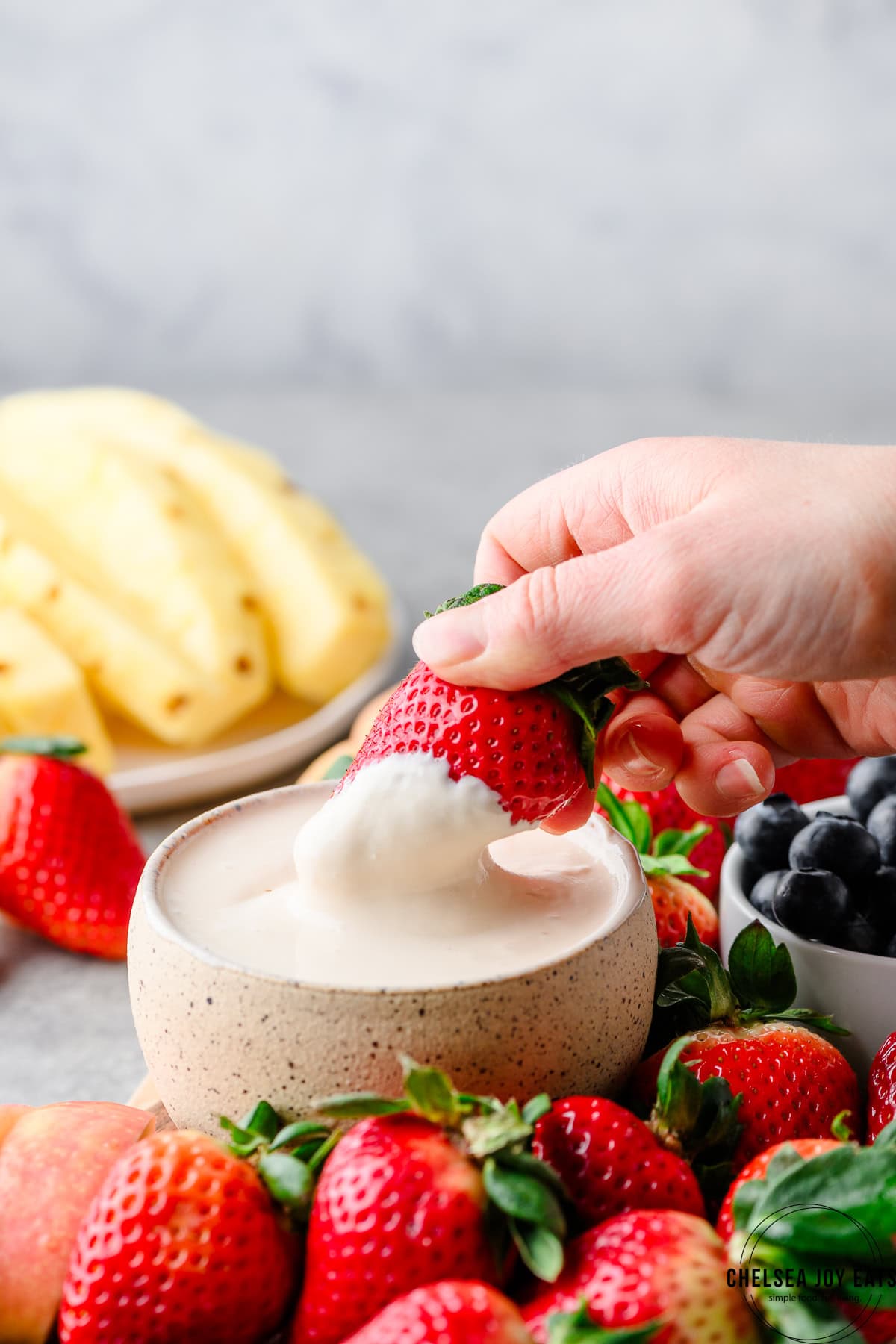 Dipping a strawberry into homemade cottage cheese fruit dip, with pineapple, more strawberries, and blueberries around the bowl of dip