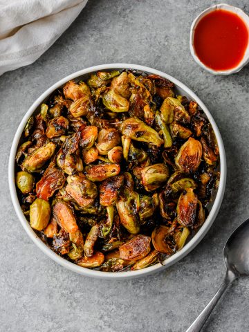 Serving bowl filled with saucy hot honey brussels sprouts with a serving spoon alongside