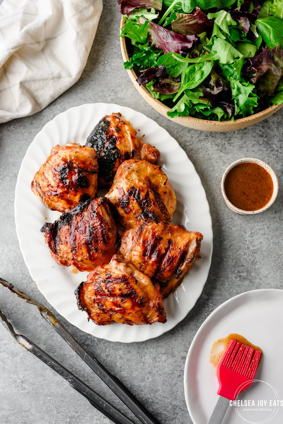 Platter of grilled chicken thighs with kitchen tongs and sauce alongside.