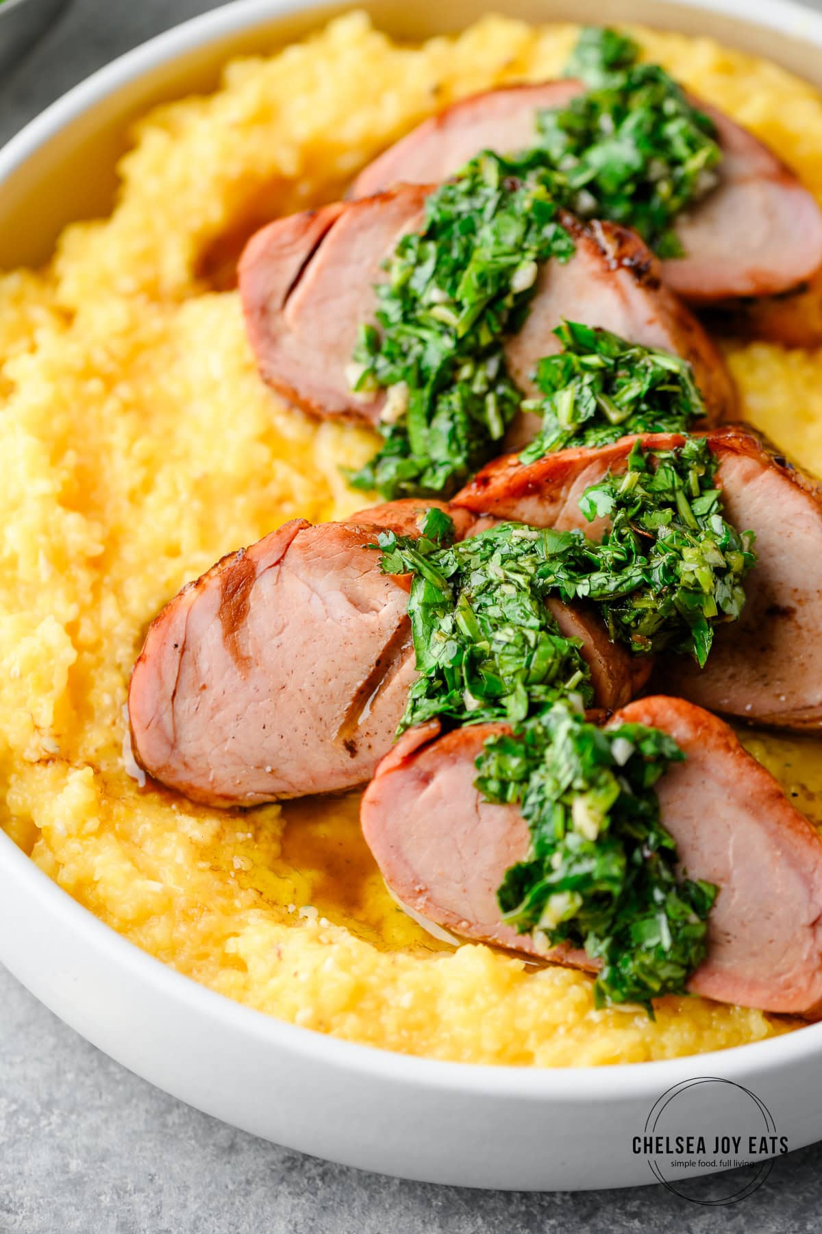 Closeup of smoked pork tenderloin coins served over polenta with fresh chimichurri drizzled overtop