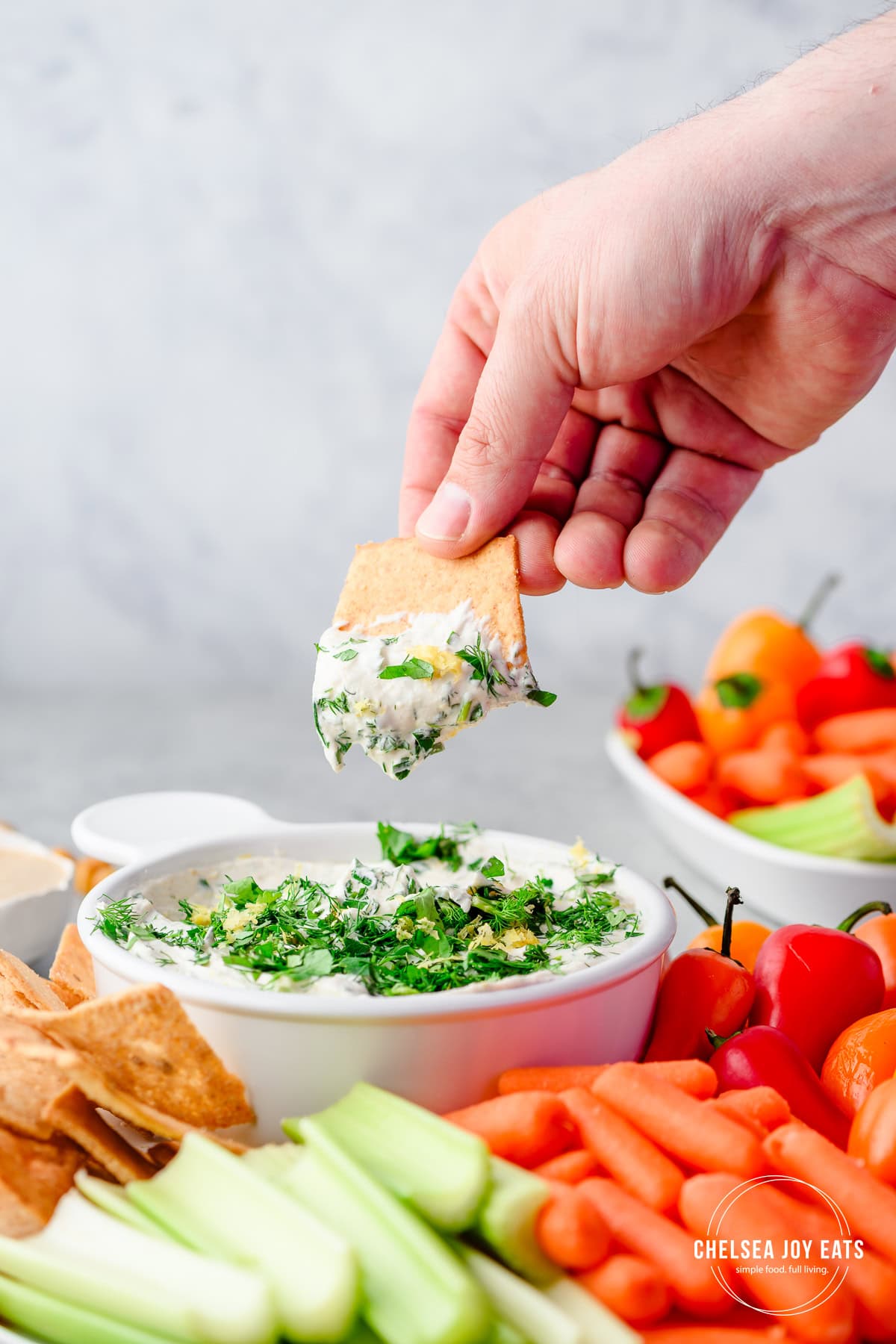 Person dipping a pita chip into an herby tahini yogurt dip with more chips and fresh vegetables all around