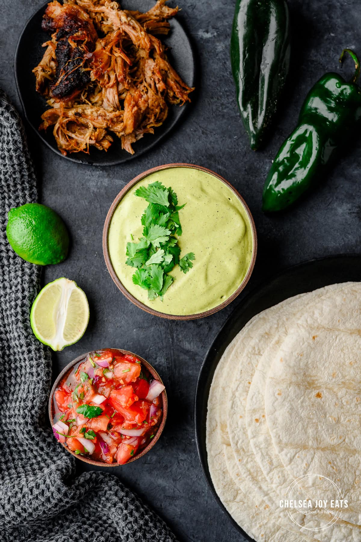Overhead view of poblano cream sauce in a bowl garnished with fresh cilantro, with ingredients for tacos like tortillas, pico de gallo, meat, and additional poblanos arranged around