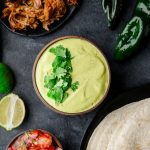 Overhead view of a bowl filled with vibrant poblano sauce made with Greek yogurt, with tortillas, meat, and fresh poblanos arranged around