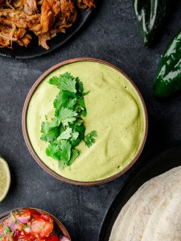 Overhead view of a bowl filled with vibrant poblano sauce made with Greek yogurt, with tortillas, meat, and fresh poblanos arranged around