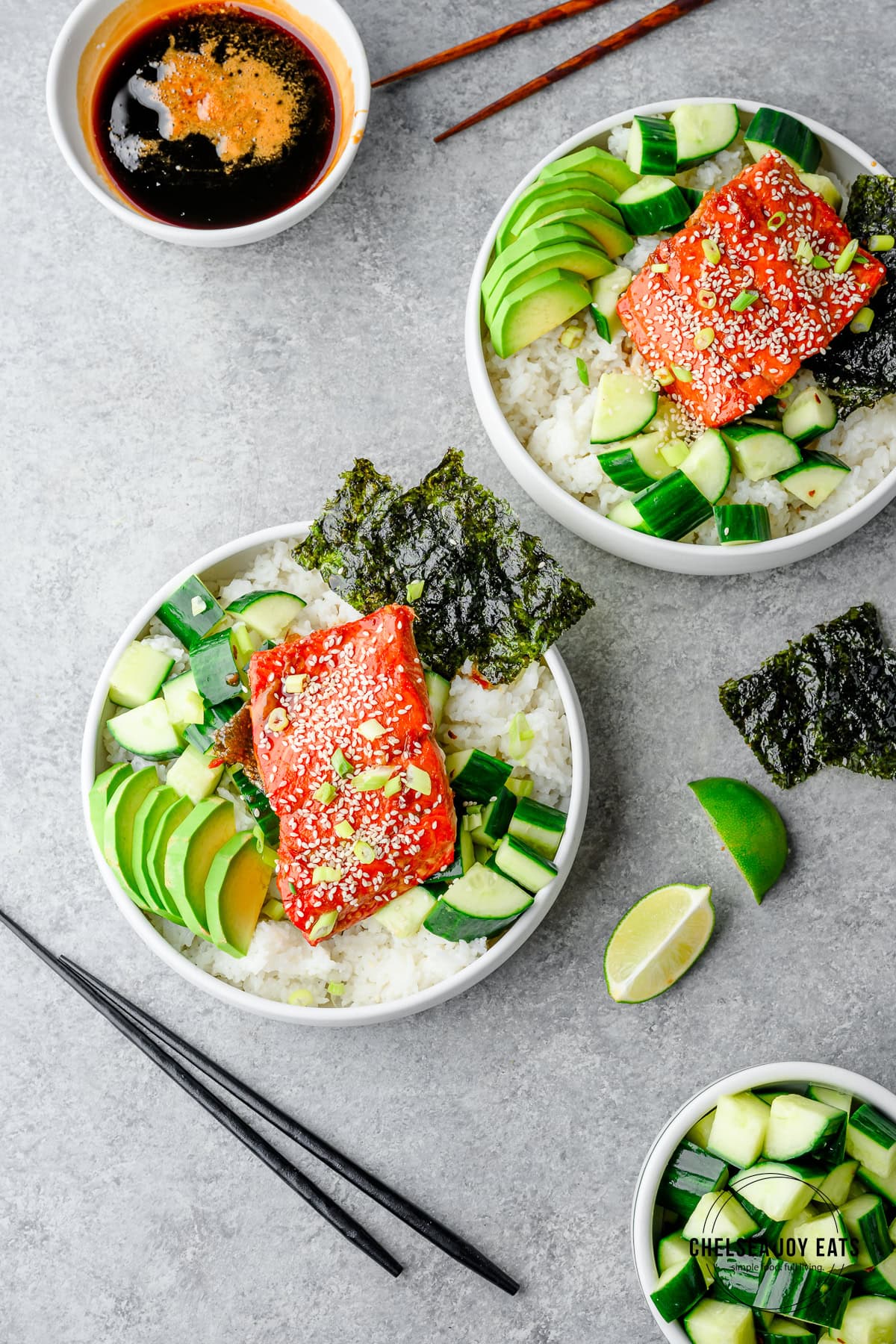 	Overhead view of rice bowls topped with cucumber salad, avocado, and teriyaki salmon with extra bowls of cucumber salad and teriyaki sauce on the side