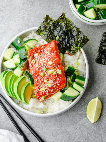 Teriyaki salmon and rice bowl served with cucumber salad, sliced avocado, and nori