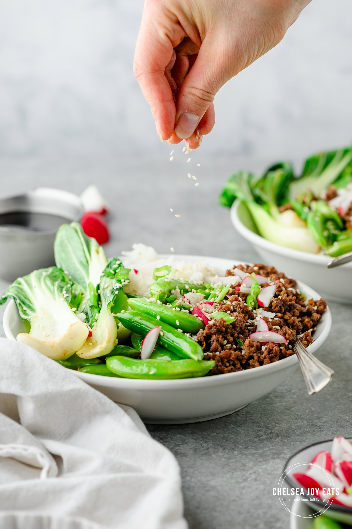 Person garnishing a bowl of veggies, rice, and ground beef with sesame seeds