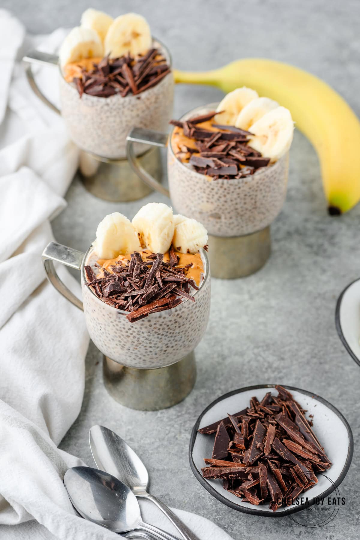 Peanut butter and banana chia pudding in individual cups topped with sliced bananas, peanut butter, and chopped chocolate
