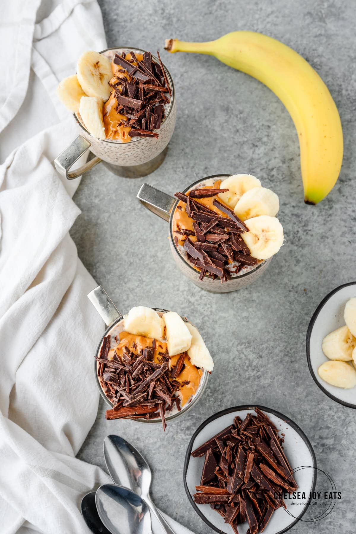 Overhead view of individual servings of banana and peanut butter chia puddings topped with chopped chocolate, banana slices, and peanut butter