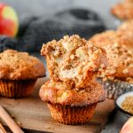 Two gluten free apple muffins stacked on top of one another, one with a bite missing, and more muffins behind