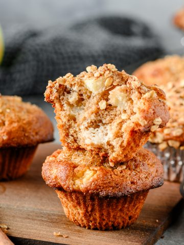 Two gluten free apple muffins stacked on top of one another, one with a bite missing, and more muffins behind