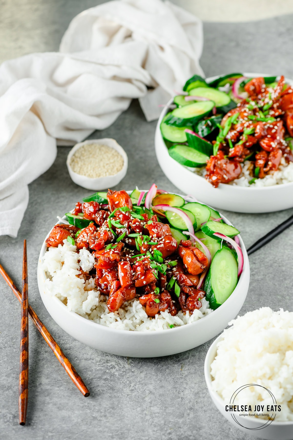 Bowls of chicken teriyaki served with rice and marinated cucumbers, with extra rice and chopsticks alongside