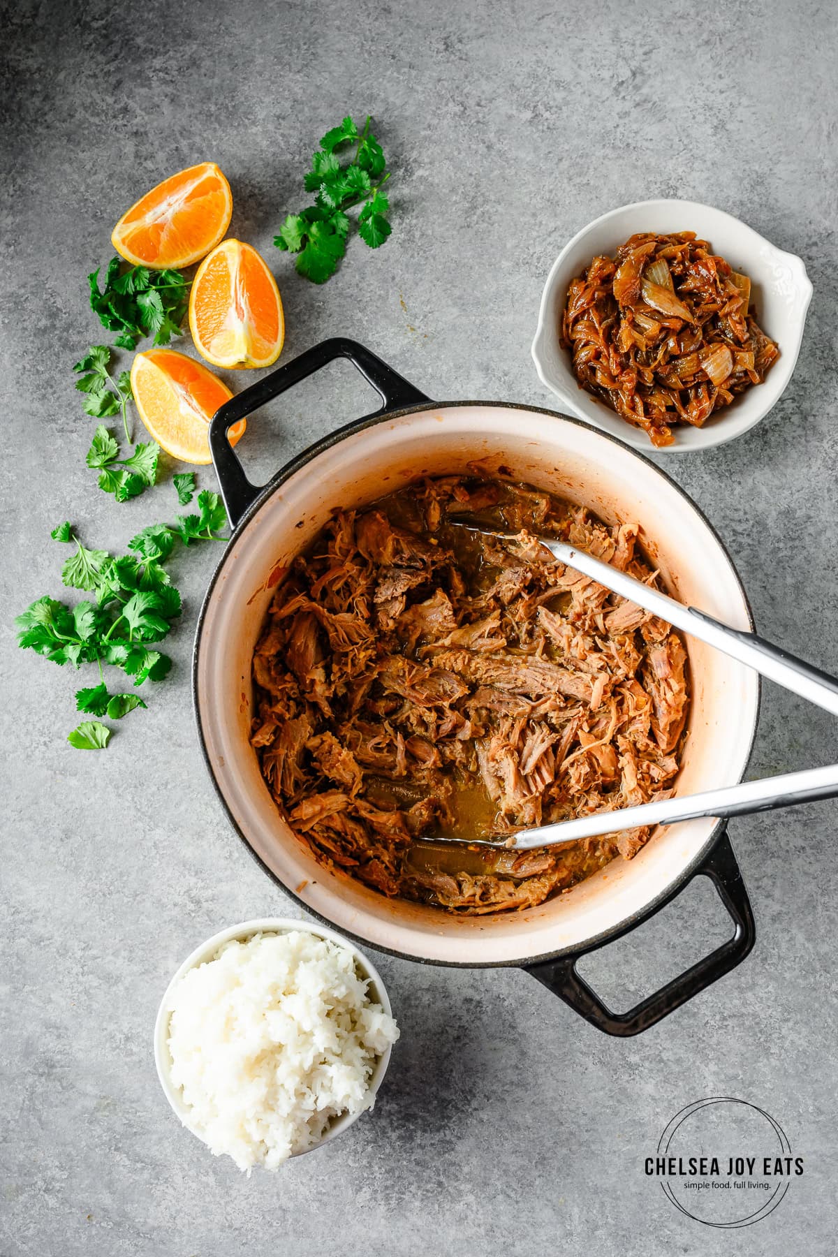 Overhead view of a Dutch oven with pulled braised pork shoulder with bowls of caramelized onion and rice alongside