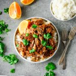 Overhead view of a bowl with braised pork shoulder served over rice, garnished with caramelized onions, cilantro, and an orange wedge.