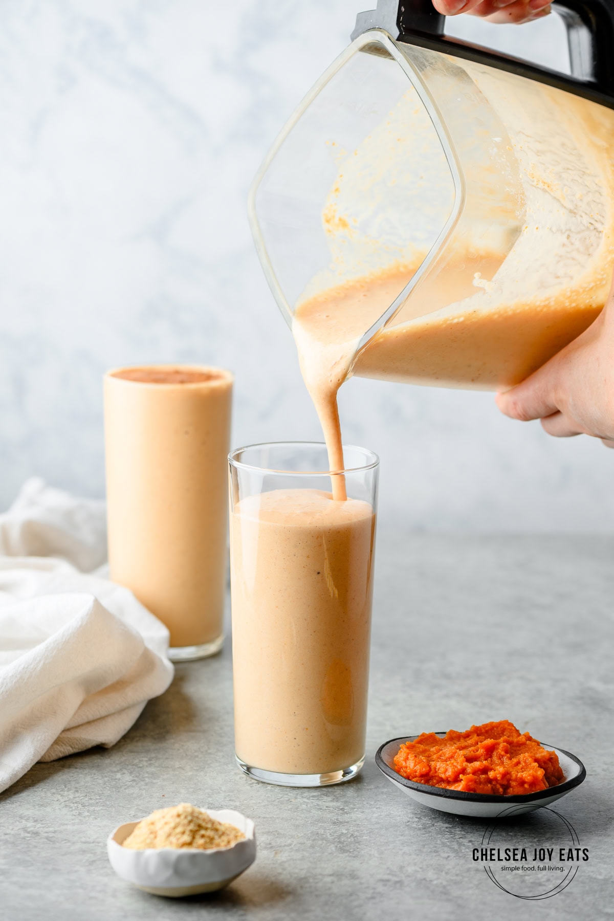 Pouring protein-packed pumpkin smoothie into tall clear glasses, with dishes of pumpkin puree and flaxseeds nearby