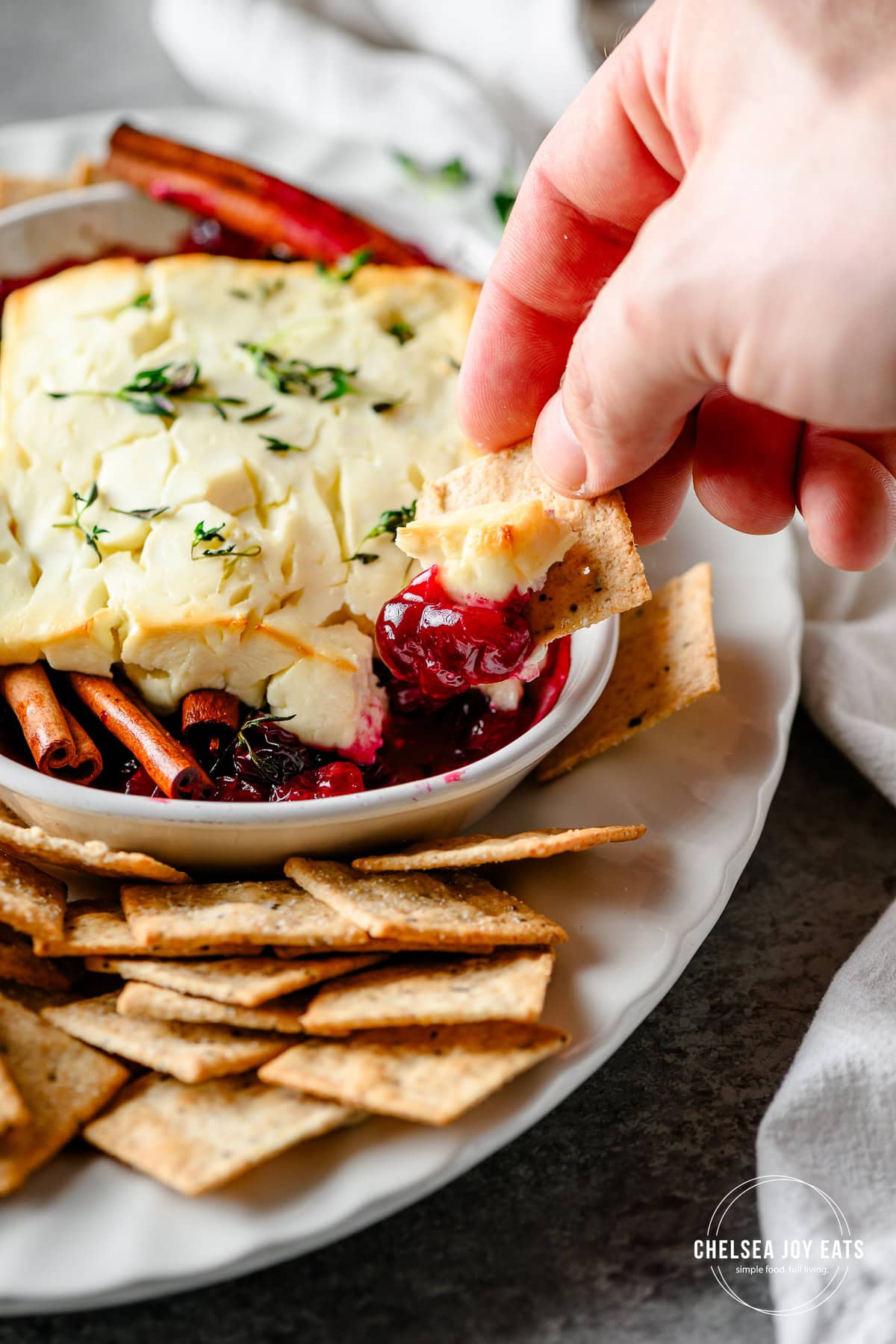 Closeup of a hand scooping baked feta cheese and cranberries out of a dish with a cracker
