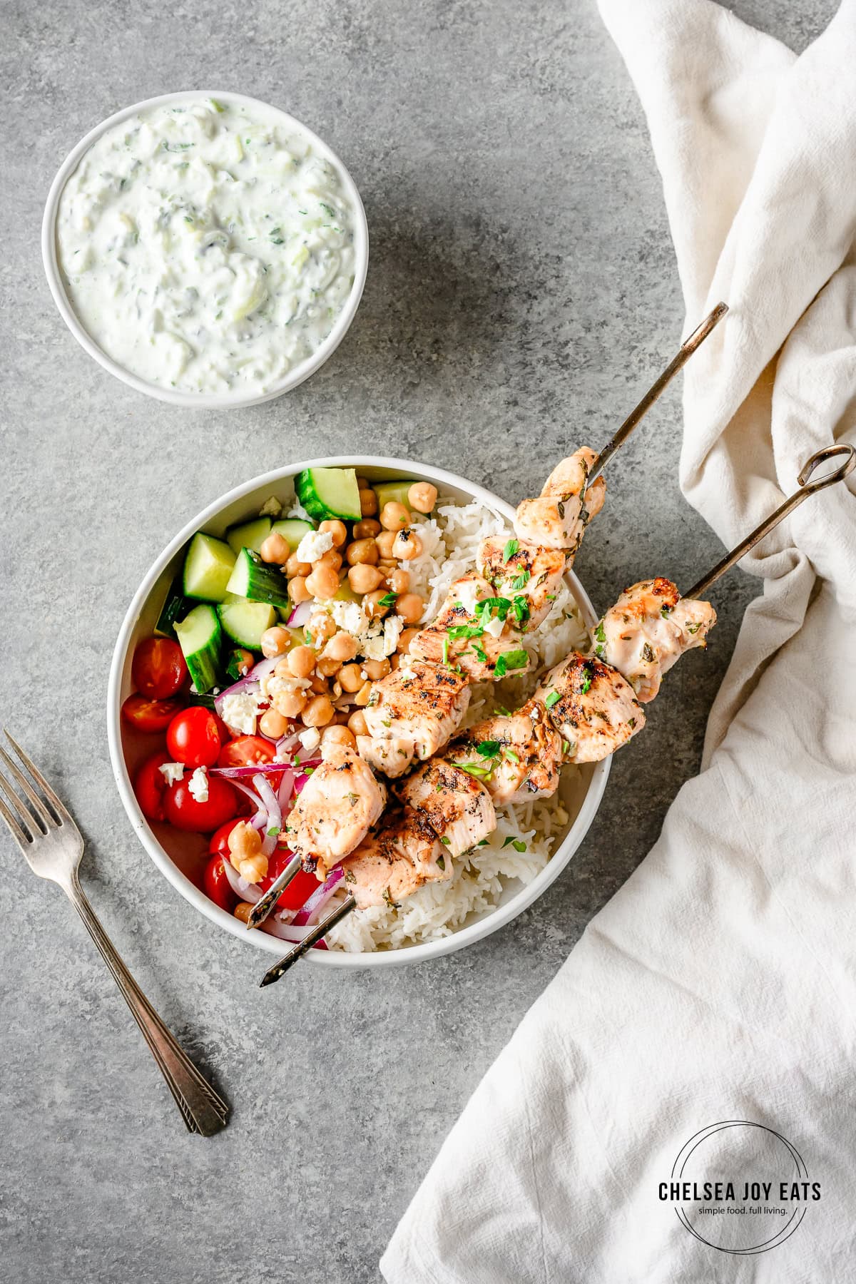 Overhead view of chicken souvlaki bowls with rice, chicken skewers, fresh veggies, and a bowl of tzatziki sauce beside