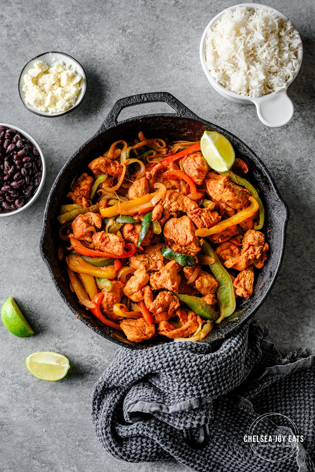 Cast iron skillet with fajita chicken, bell peppers, and onions, surrounded by dishes of rice, cheese, and beans