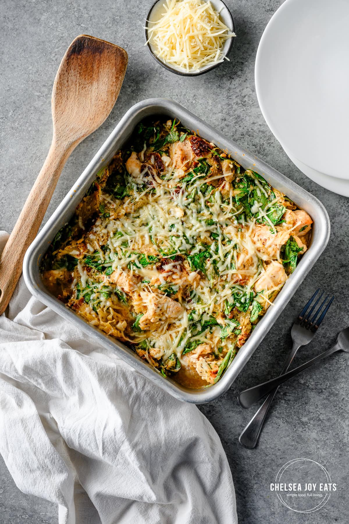 spaghetti squash casserole with feta, spinach, and chicken in a aluminum baking dish with grated Parmesan and a wooden spoon.