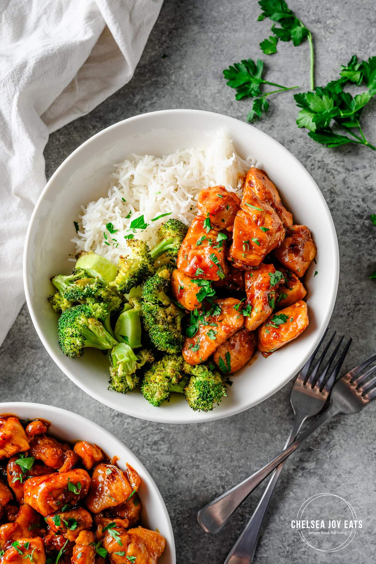 Bowl with rice, roasted broccoli, and gluten free honey garlic chicken, with forks and another bowl of chicken alongside.