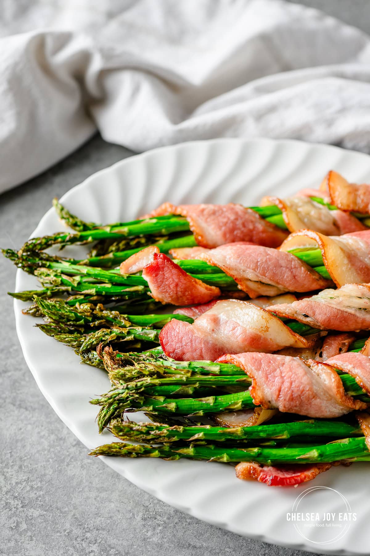 Bundles of oven-roasted bacon-wrapped asparagus piled on a white ceramic platter.