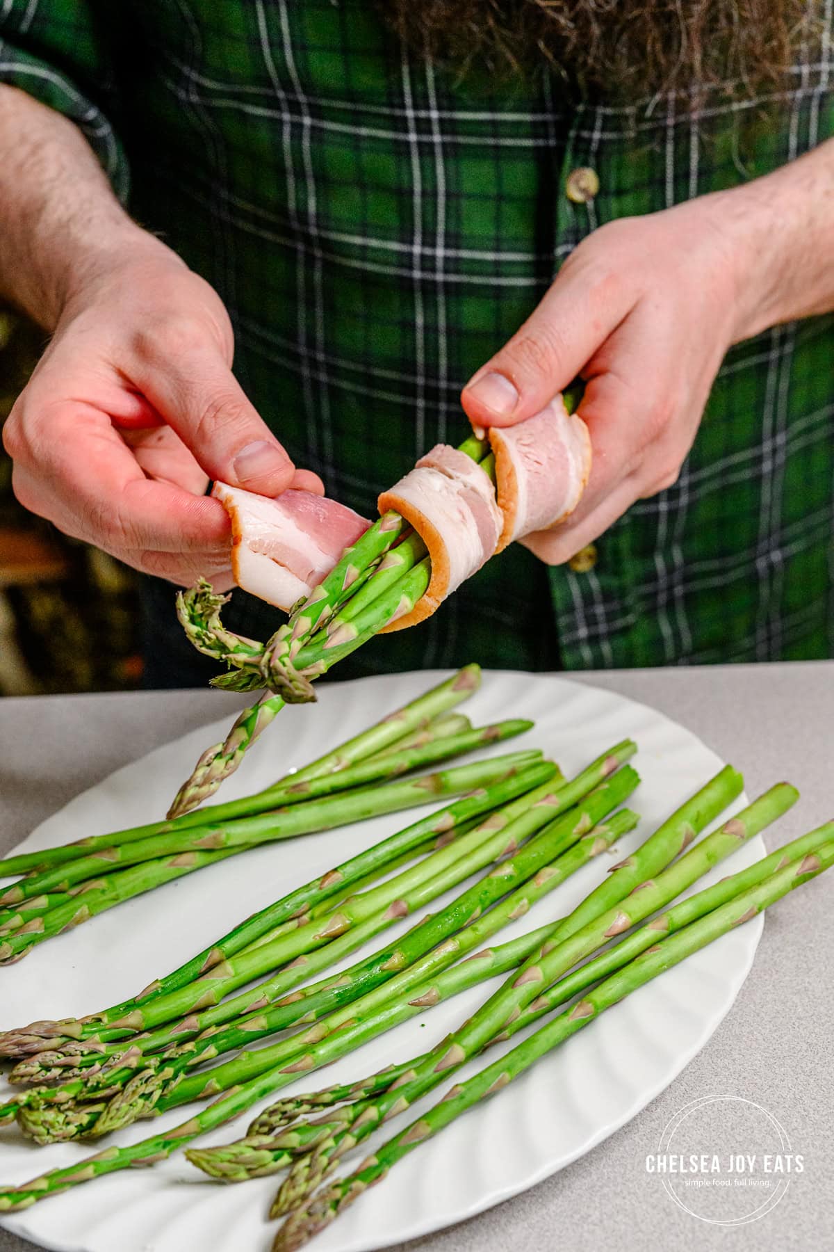 Person wrapping a bundle of 3 fresh asparagus stalks with a strip of bacon.