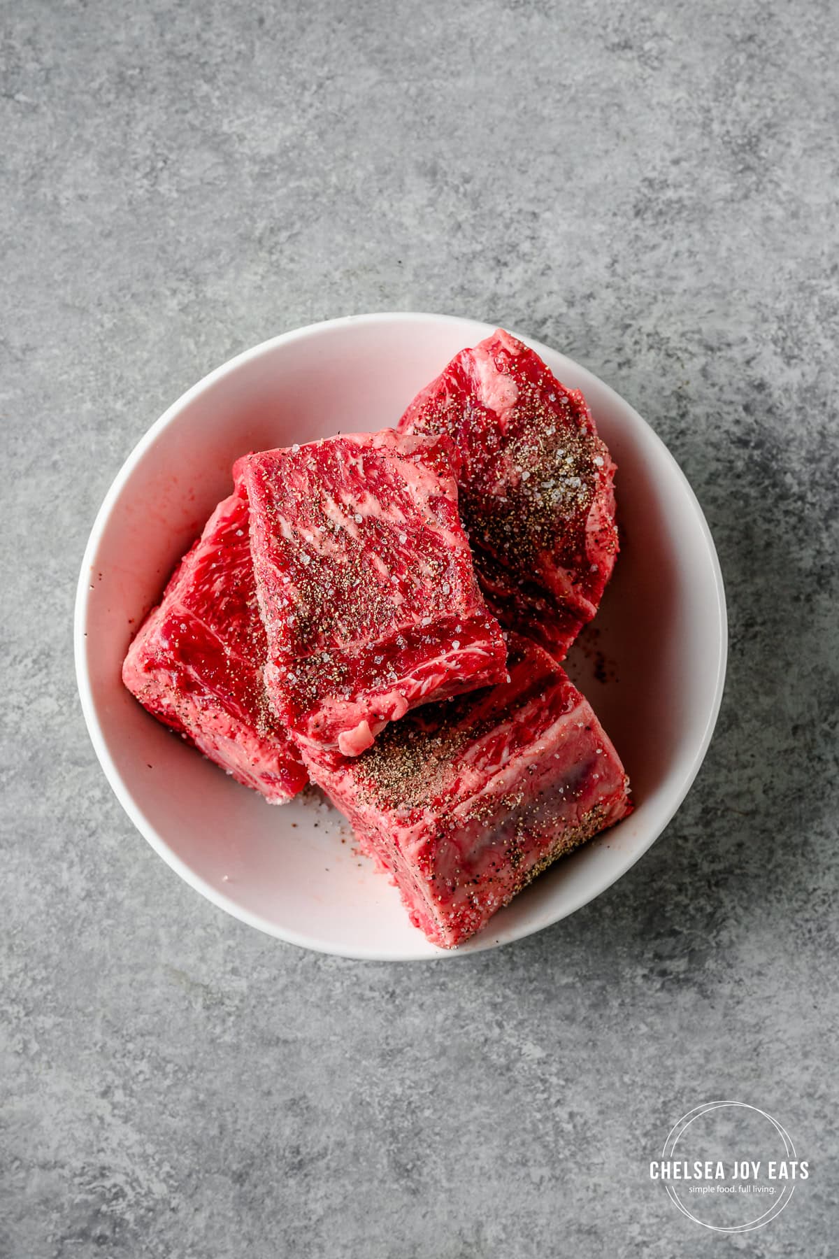 Raw beef short ribs seasoned with salt and pepper in a white bowl.