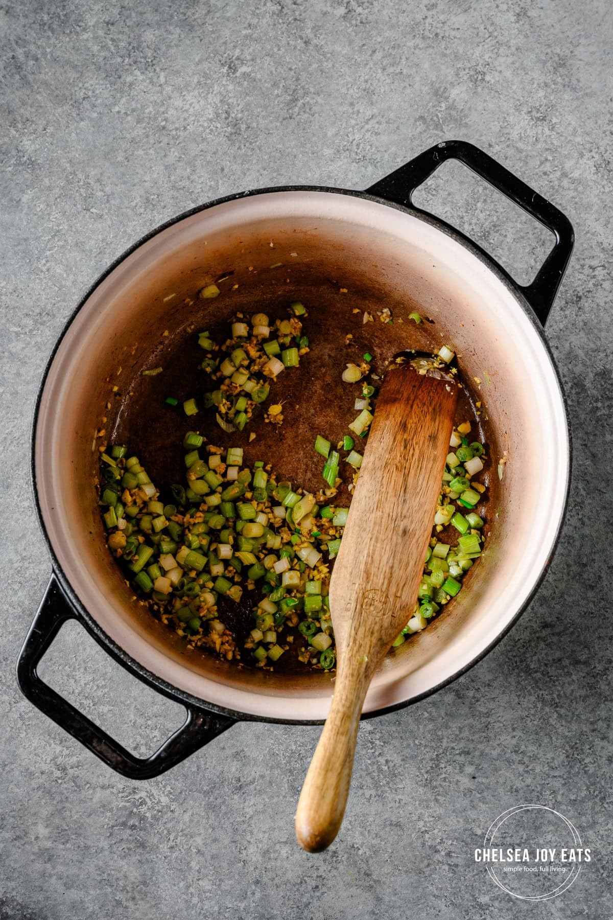 Ginger and scallion cooking in a Dutch oven with a wooden spatula stirring.