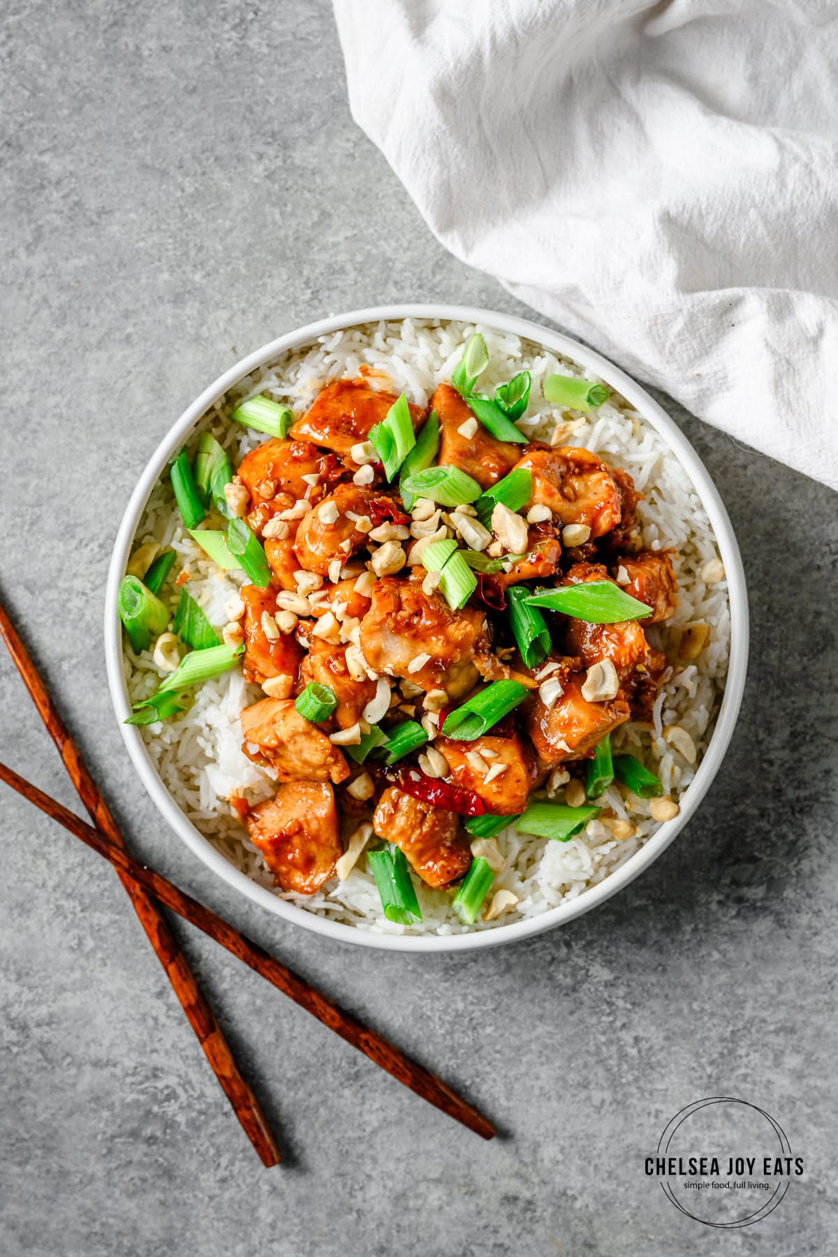 Bowl with rice and gluten free kung pao chicken topped with green onions and cashews, with a pair of chopsticks alongside.