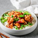 White bowl filled with rice and gluten free kung pao chicken topped with scallions and cashews.