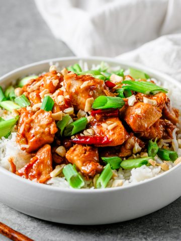 White bowl filled with rice and gluten free kung pao chicken topped with scallions and cashews.