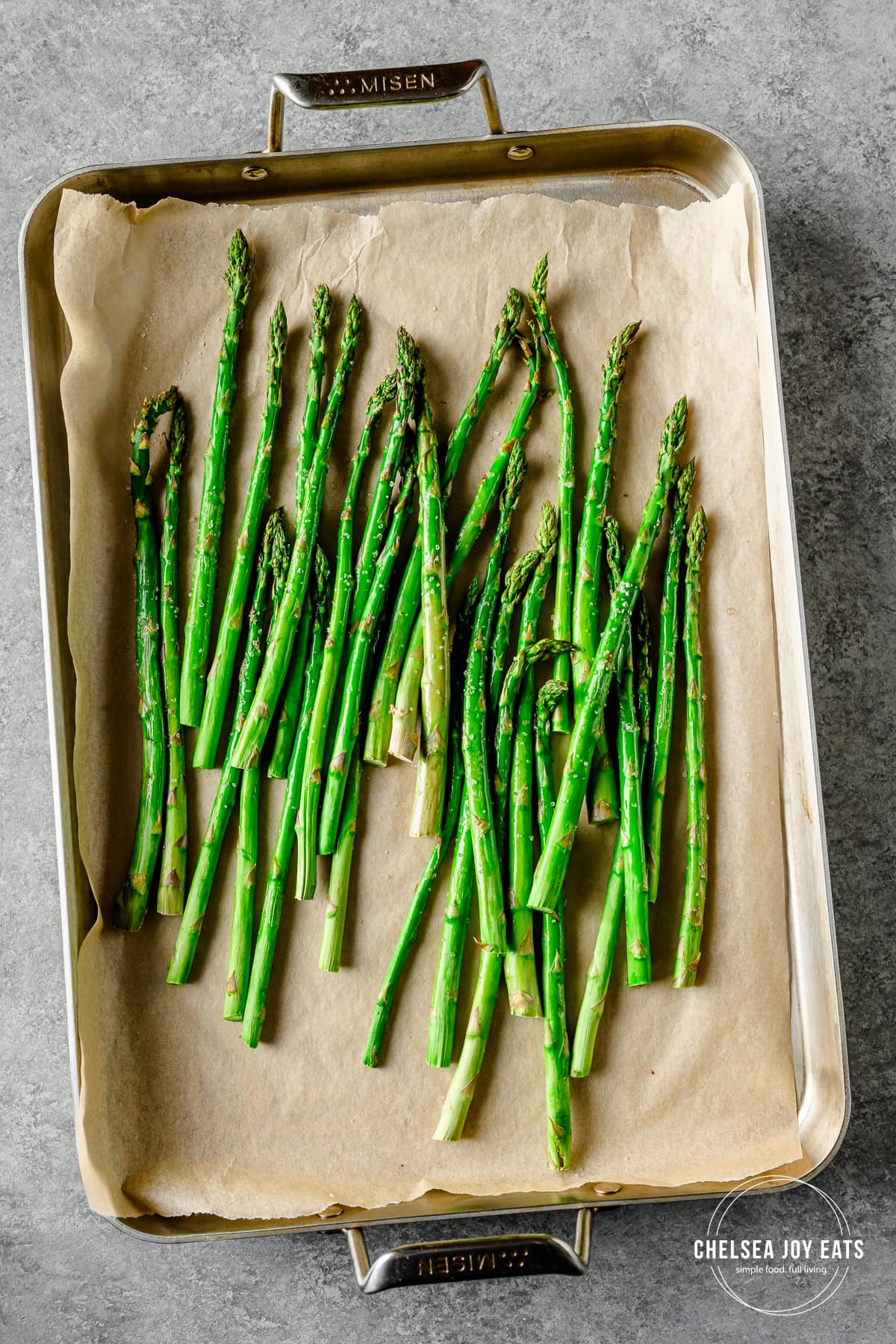 Asparagus tossed in bacon fat on a stainless steel baking sheet lined with parchment paper.