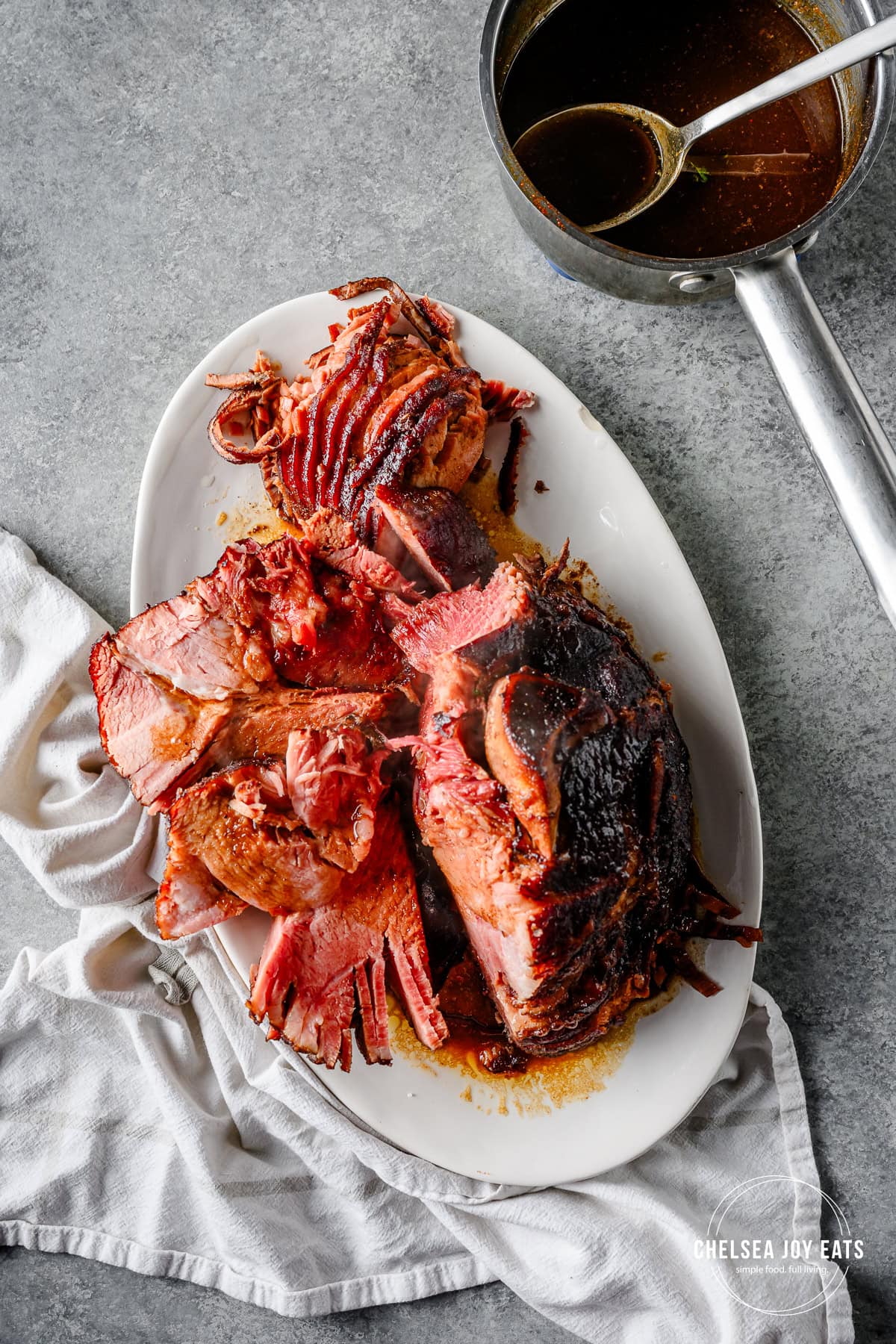 Glazed cooked ham served on a platter, half-sliced and ready to serve, with a saucepan of gluten free glaze alongside.