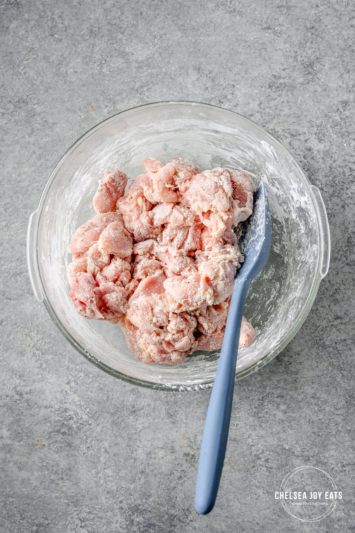 Chicken thigh pieces tossed with arrowroot flour in a bowl, with a silicon spatula mixing.
