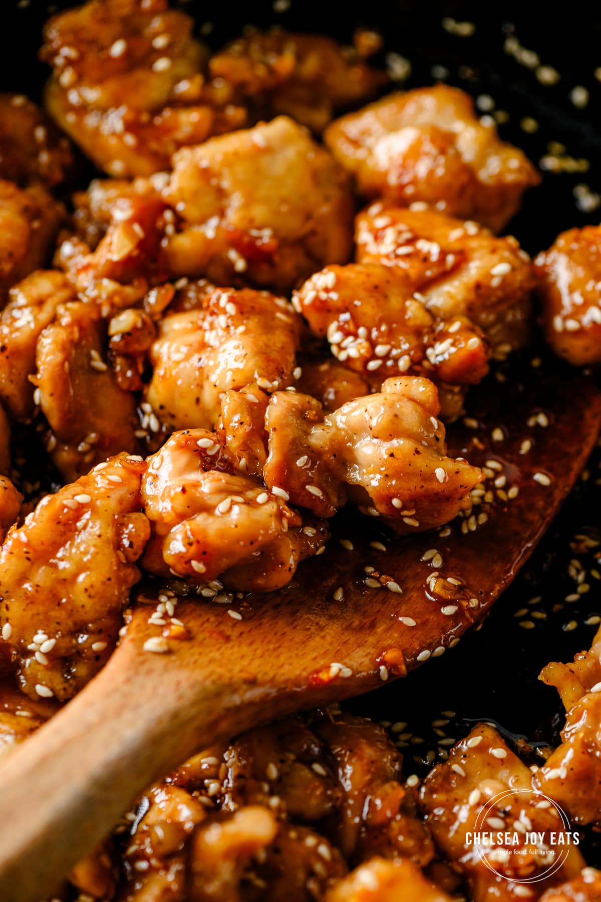 Wooden spatula stirring pieces of gluten free honey sesame chicken in a cast iron skillet.