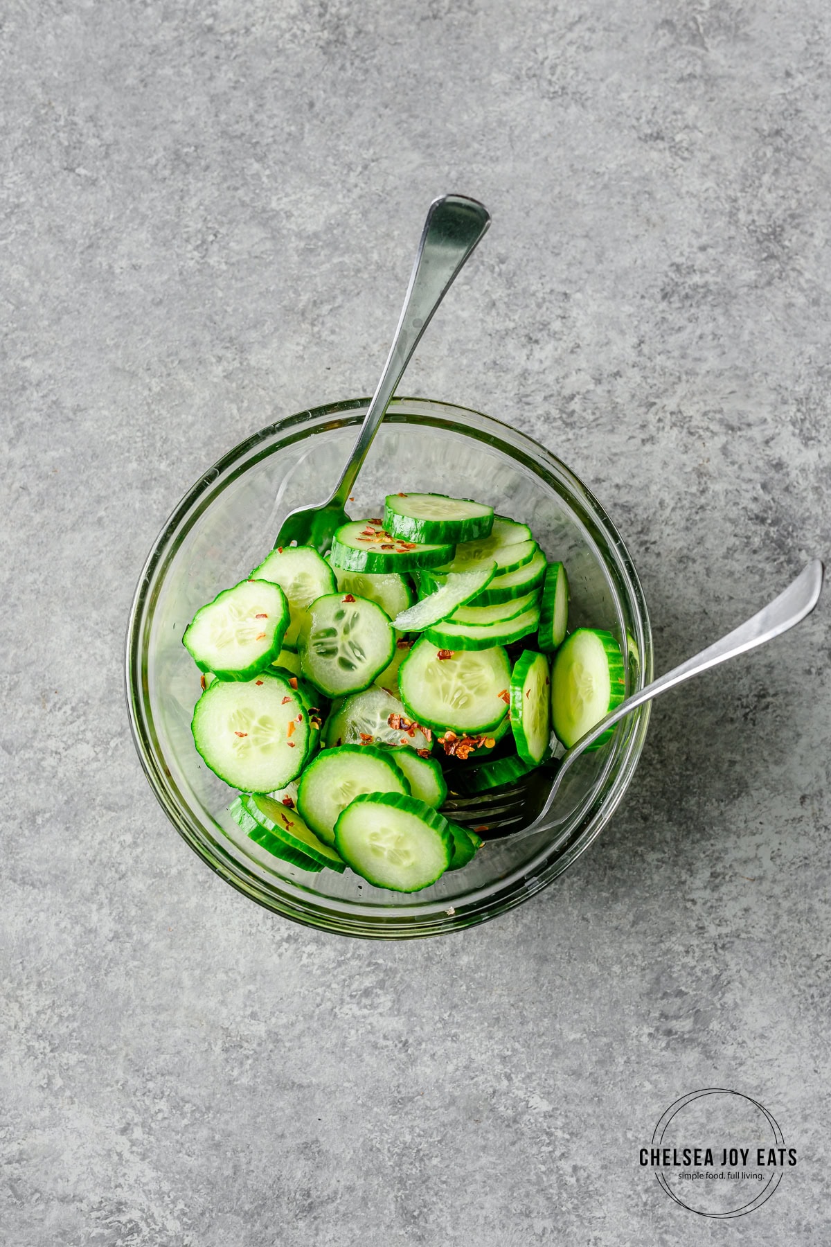 Sliced cucumbers tossed with rice vinegar and red pepper flakes in a glass bowl with forks to toss.