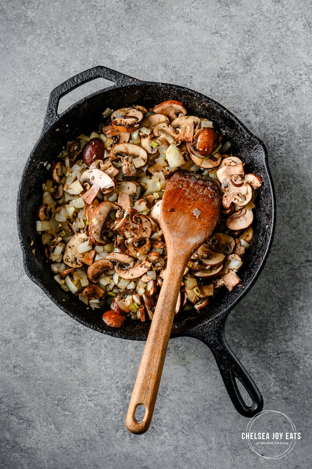 Sauteed onions and cremini mushrooms in a cast iron skillet with a wooden spatula.