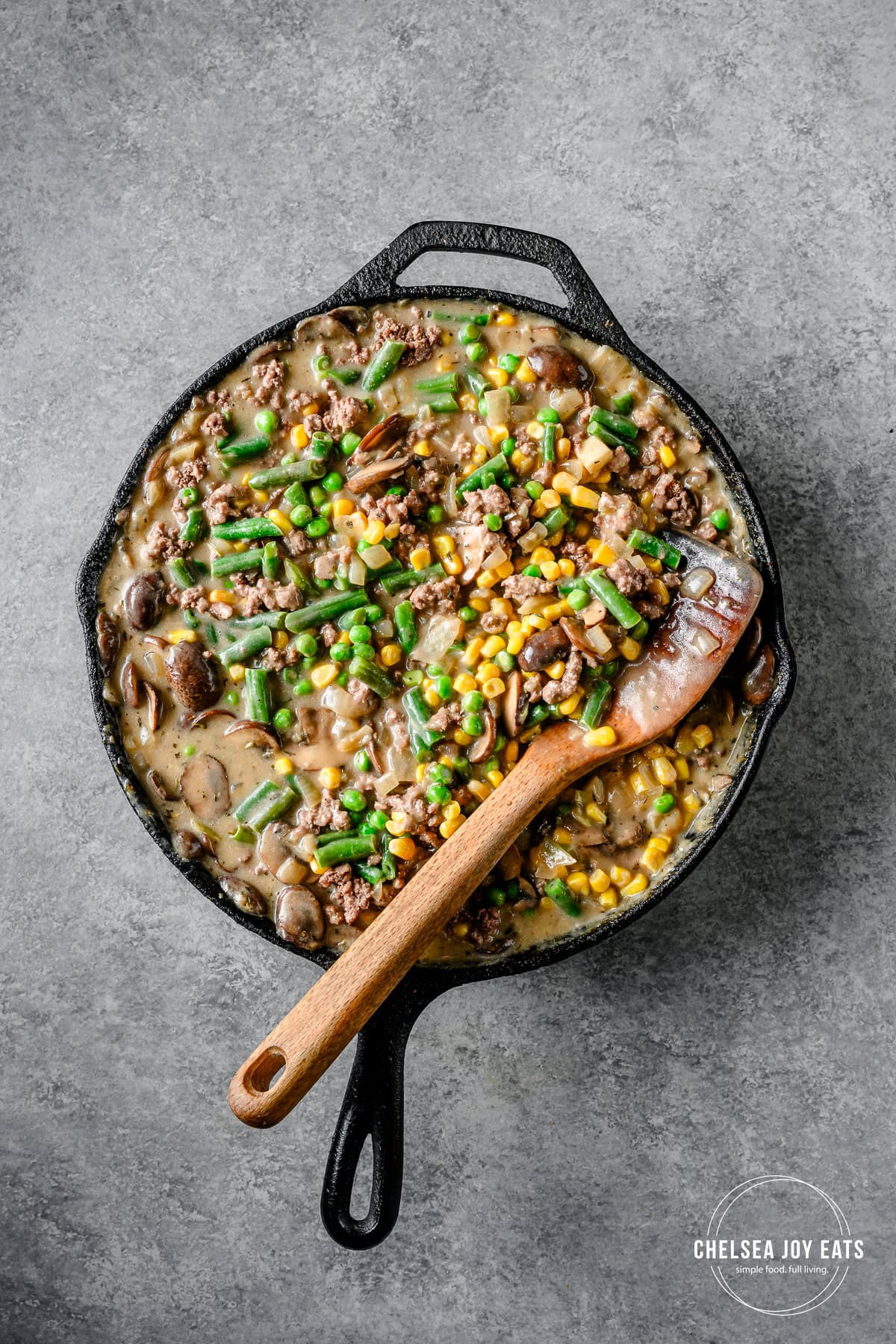 Frozen green beans, corn, and peas cooking with ground beef in a mushroom gravy in a cast iron skillet.