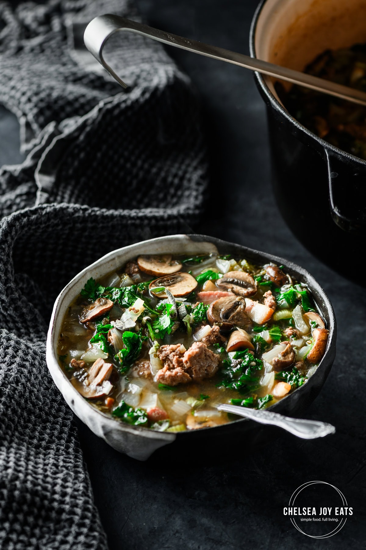 Bowl of bacon bison mushroom soup with kale and a spoon in the soup with a Dutch oven with more soup in the background.