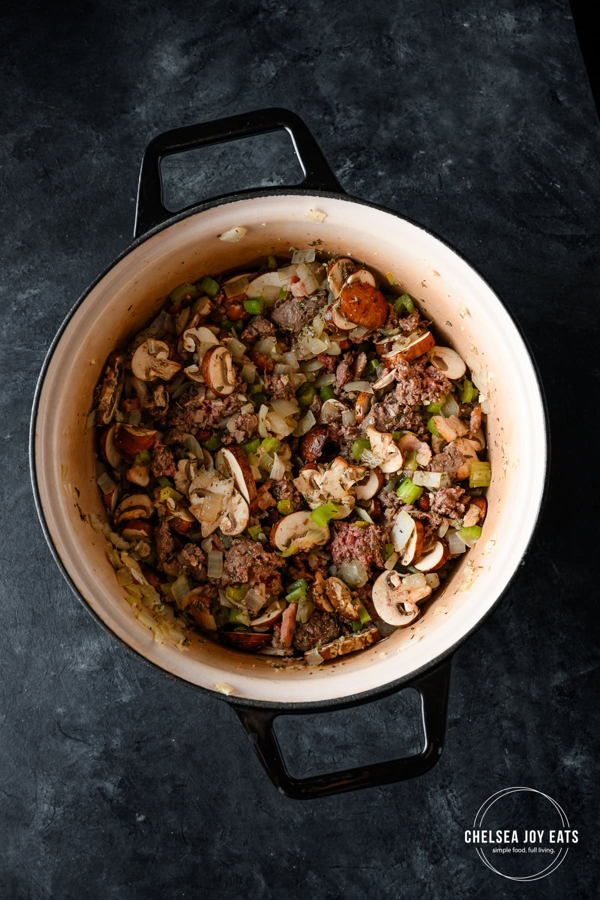 Ground bison, mushrooms, celery, onion, and herbs cooking in a Dutch oven on a dark background.
