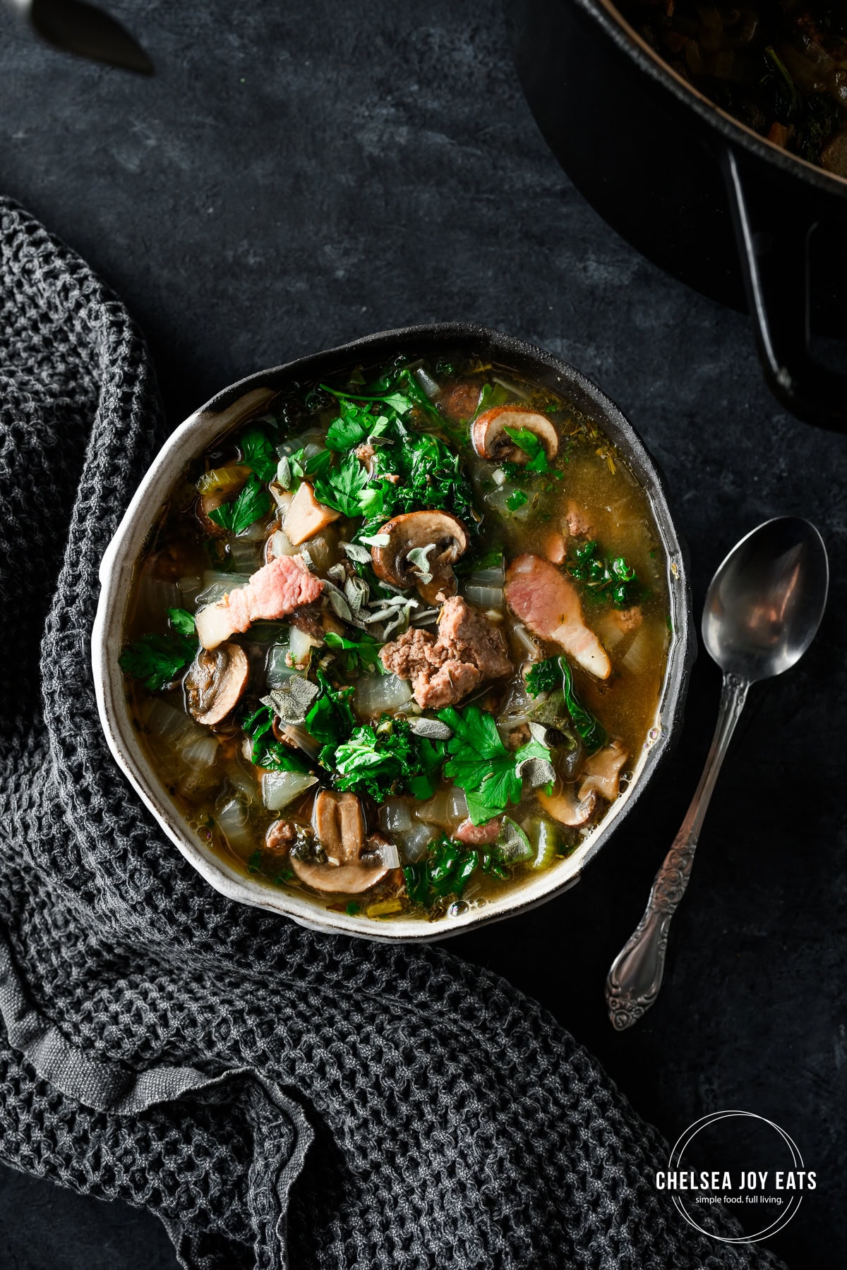 Bowl of Bacon Bison Mushroom Soup with kale on a dark counter with a spoon alongside.