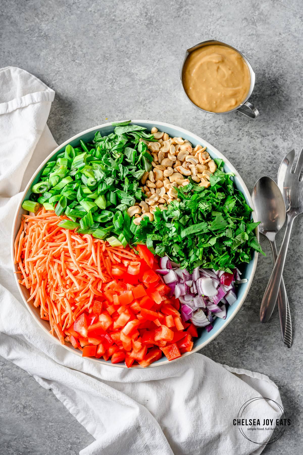 Ingredients for Thai cabbage salad assembled in a bowl with a cup of peanut dressing and serving utensils alongside.