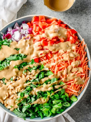 Gluten free Thai cabbage salad served in a large ceramic bowl with peanut dressing drizzled overtop, with additional dressing in a dish and serving utensils alongside.